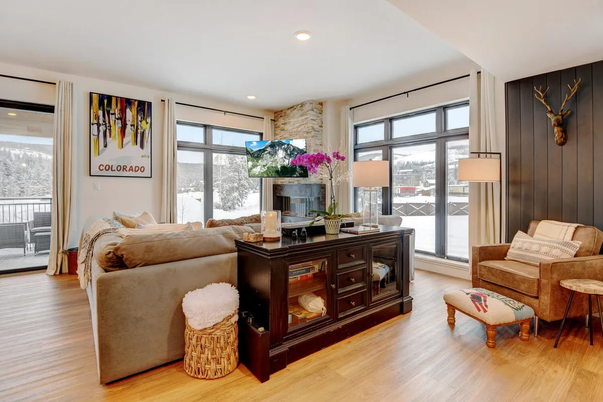 The image showcases a cozy, tastefully decorated living room with a plush sofa, stylish coffee table, warming fireplace, and a large television. Elegant artwork and a fresh flower arrangement create a relaxing and inviting atmosphere, perfect for gathering with family or friends after a day of mountain adventures. Large windows let in natural light, hinting at beautiful snowy views outside.