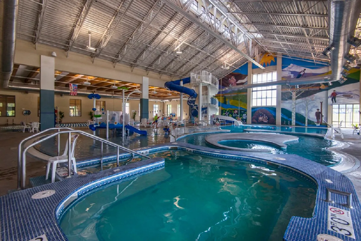 An energetic indoor swimming facility with water slides, a swimming pool, a colorful mural, and children playing. The mood is joyful and lively, showing a recreational space ideal for families and group fun.