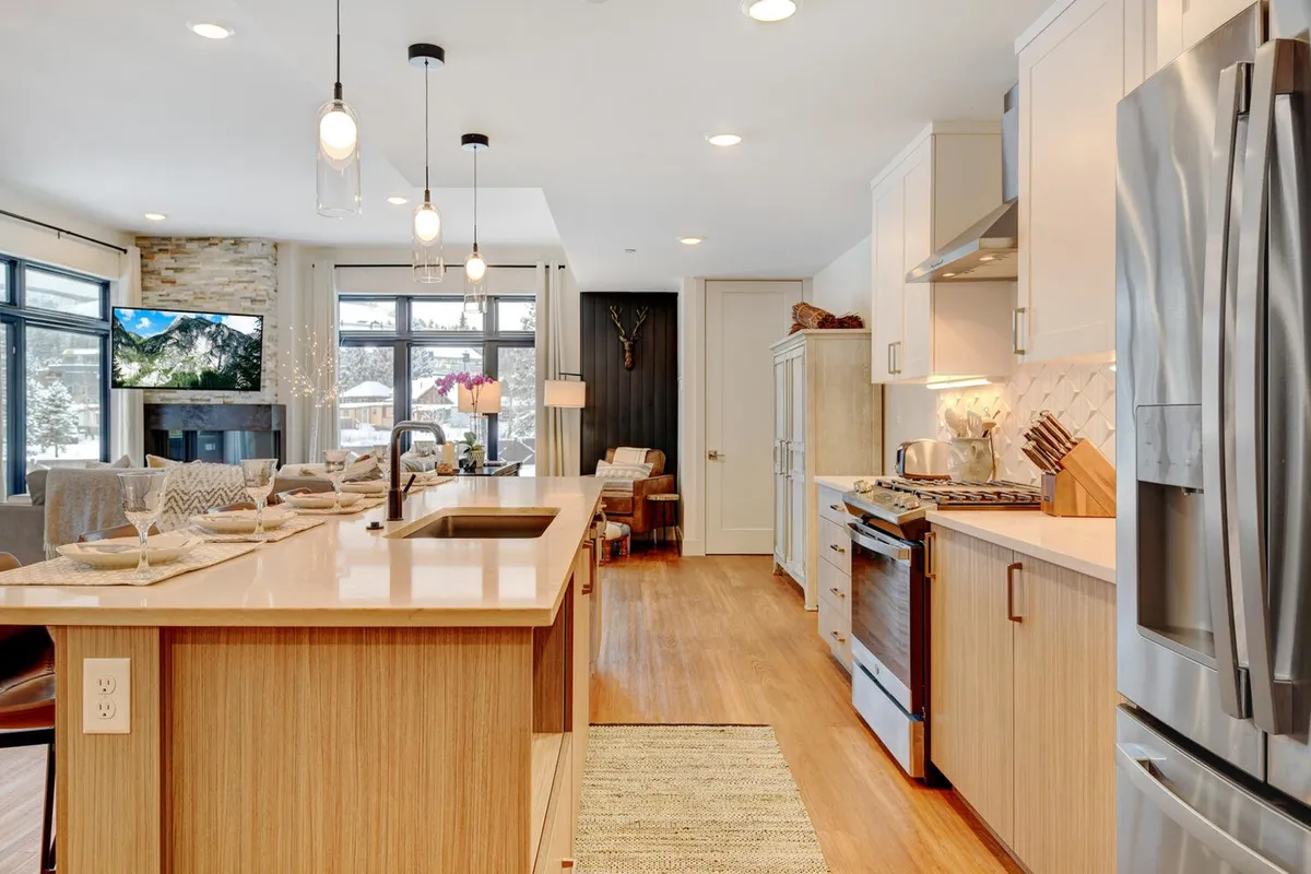 This image captures a spacious, modern open-concept area featuring the kitchen island with bar stools, stainless steel appliances, a sleek fireplace, large windows with mountain scenery, and decorative lighting. The scene emphasizes a communal vibe ideal for entertaining, cooking, and relaxing together.