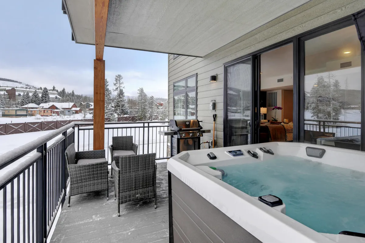 This image shows a private balcony outfitted with a hot tub, outdoor seating, and a grill, all facing a scenic snowy mountain backdrop. The area is clearly intended for relaxation and outdoor dining, with the hot tub bubbling invitingly and a grill ready for evening cookouts. The snow-covered landscape beyond enhances the sense of alpine retreat.