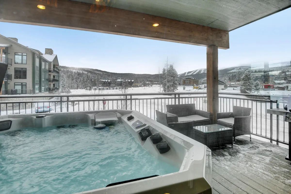 A cozy hot tub sits on a snow-fringed balcony, surrounded by inviting outdoor seating and overlooking a beautiful sunrise or sunset over the mountains. The snow creates a tranquil winter atmosphere, while the seating suggests the space is ideal for both quiet solitude and small gatherings under the stars.