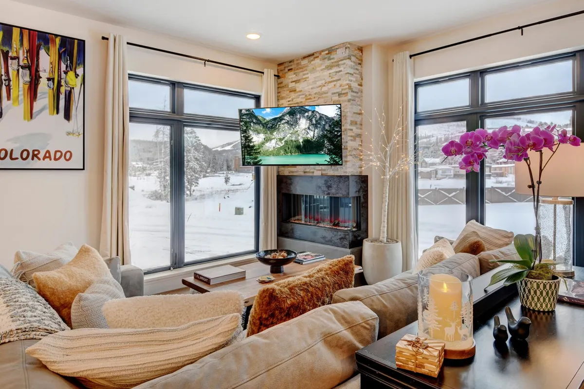 A welcoming living room scene with a comfortable sofa, floor-to-ceiling windows showcasing snowy mountain views, a modern color-changing fireplace, decorative art, and a TV displaying nature imagery. The setting is perfect for family relaxation, movie nights, or après-ski unwinding.