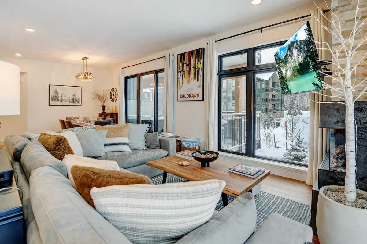 The image captures a cozy living room with a comfortable sofa adorned with decorative pillows, a coffee table, and large windows framing a snowy mountain landscape outside. A fireplace adds a sense of warmth and relaxation to the space, making it inviting after a day of outdoor adventure.