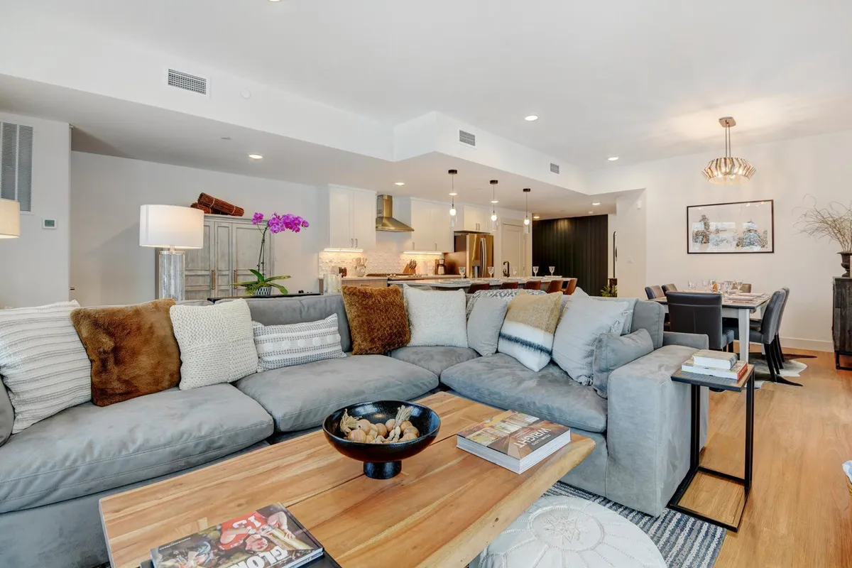 This image offers a broad perspective of an open-concept living area, showing a large sectional sofa, a wooden coffee table, a dining area, and a modern kitchen in the background. The space is bright and inviting, suggesting opportunities for relaxation and socializing after exploring local trails or slopes.
