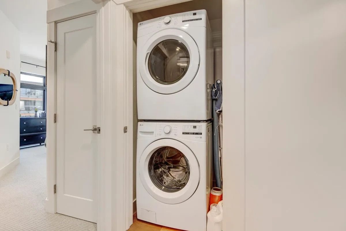 A bright and tidy laundry area featuring a stacked washer and dryer, with visible cleaning supplies neatly arranged nearby. The space exudes organization and cleanliness, emphasizing practical upkeep and a well-maintained home environment.