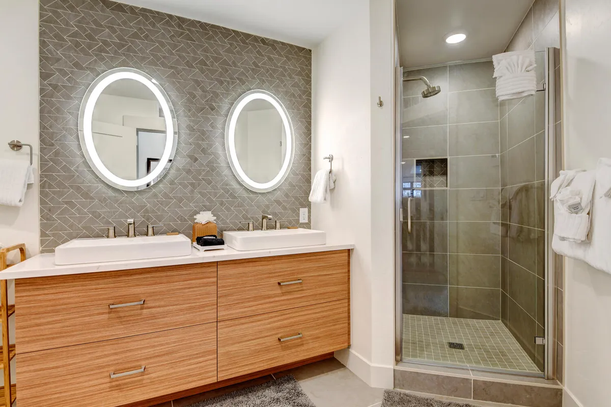 A sophisticated bathroom with double sinks, oval mirrors featuring backlighting, textured wall tiles, and a glass-enclosed walk-in shower. The space exudes a spa-like ambiance, perfect for relaxation and self-care routines.