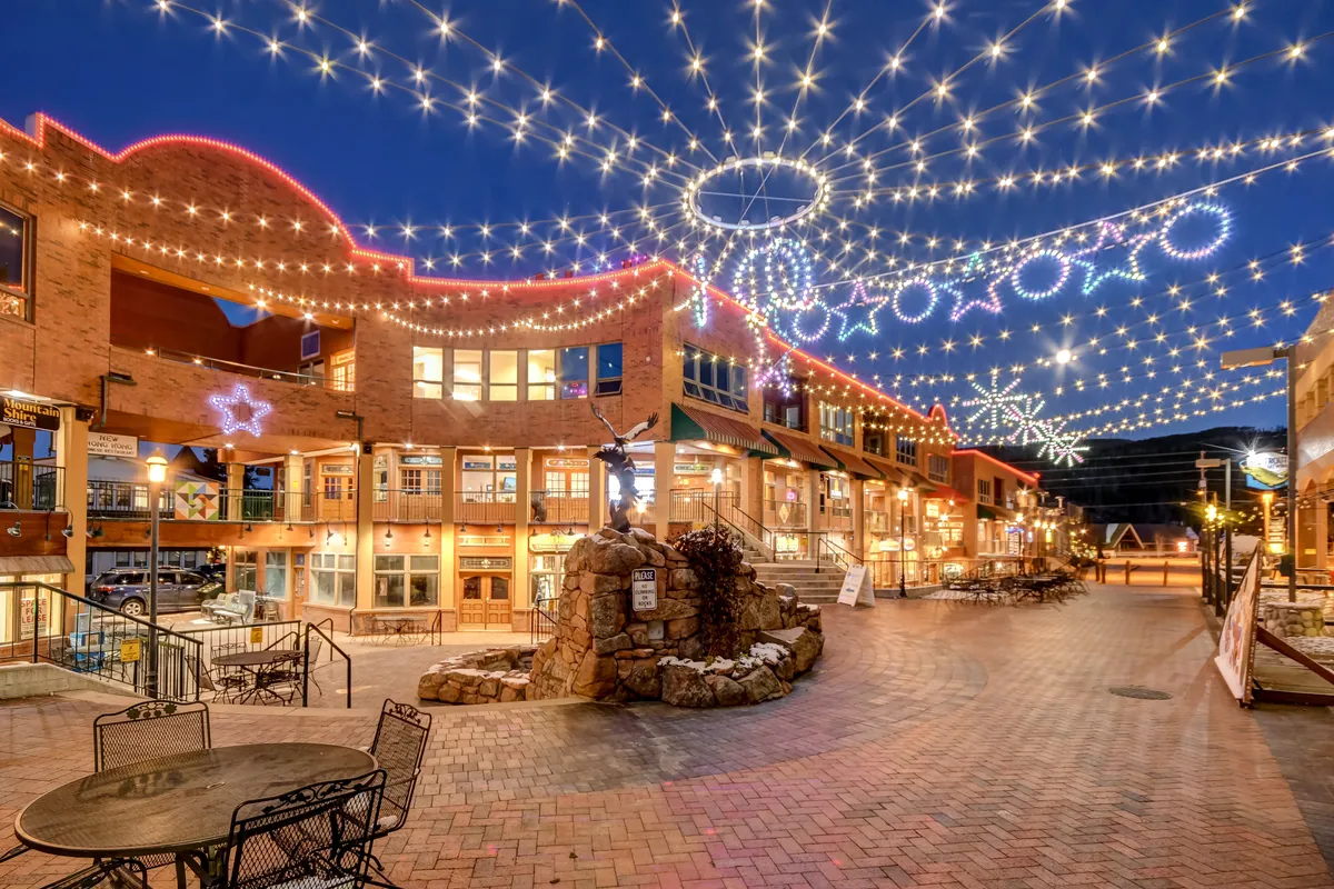 A charming outdoor plaza with string lights overhead, a brick pathway, a decorative stone fountain, and shops with large windows. Groups of people stroll and socialize, emphasizing the festive, walkable downtown experience.