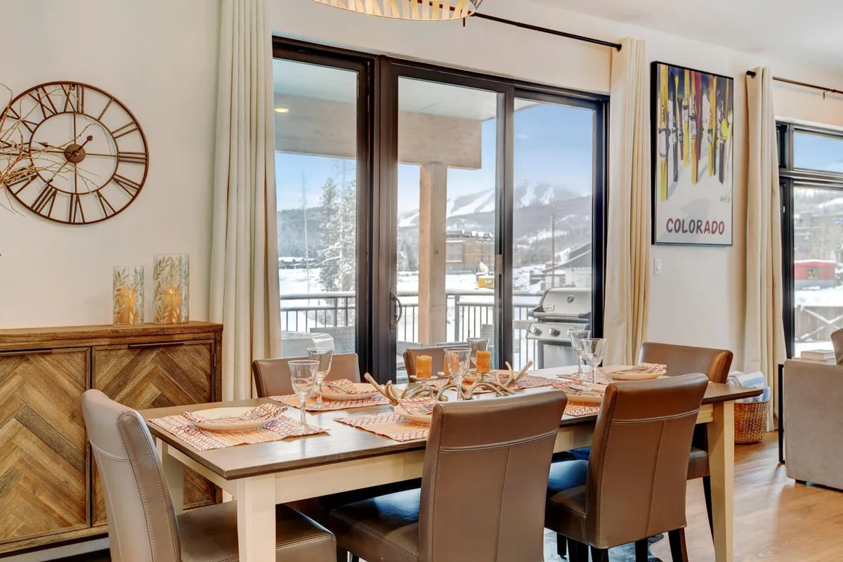 A dining area set for a meal, with a large table ready for group gatherings, big windows opening to mountain views, and tasteful wall art. The mood is warm and inviting, perfect for sharing meals and family stories.