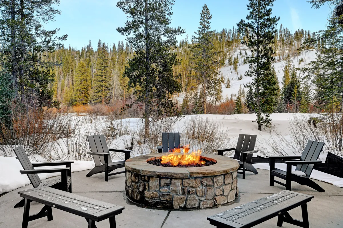 A communal firepit with surrounding Adirondack chairs sits amidst a snowy landscape and pine trees, offering a warm and welcoming spot for guests to gather, share stories, and enjoy hot beverages. The view of the fire against the snow highlights the communal and social opportunities available outside the private balcony.
