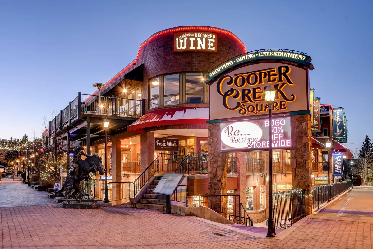 A lively street scene featuring the Cooper Creek Square sign, a wine shop sign, inviting outdoor seating areas, and decorative lighting. People are visible enjoying shopping, dining, and socializing, creating a vibrant, bustling downtown atmosphere.