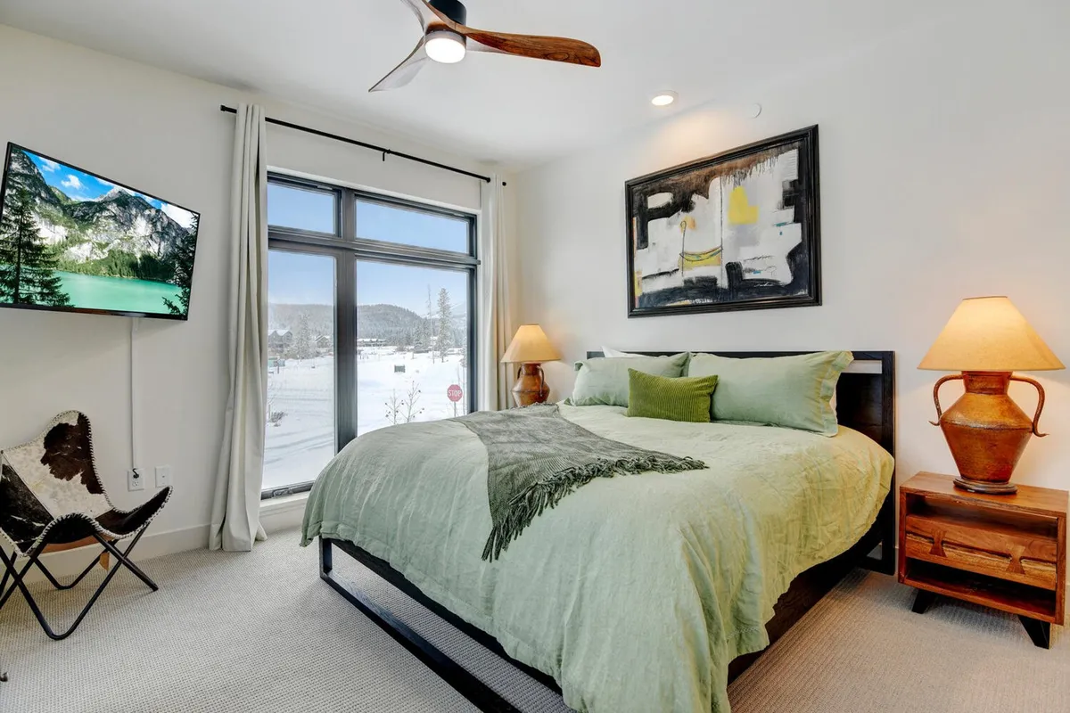 A serene bedroom featuring a plush bed with soft green bedding, large decorative artwork above the headboard, a TV mounted on the wall, and wooden bedside tables with lamps. The space is designed for relaxation, with a soothing color palette and gentle lighting.