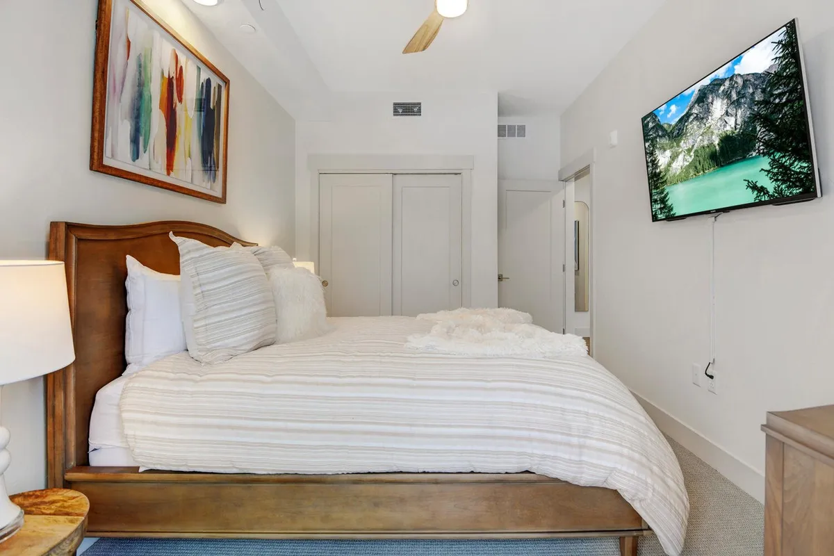 A serene bedroom with fresh white bedding, warm wood accents, a lamp for soft lighting, and a wall-mounted TV showing a scenic landscape. Everything appears immaculate and inviting, emphasizing comfort and tranquility.