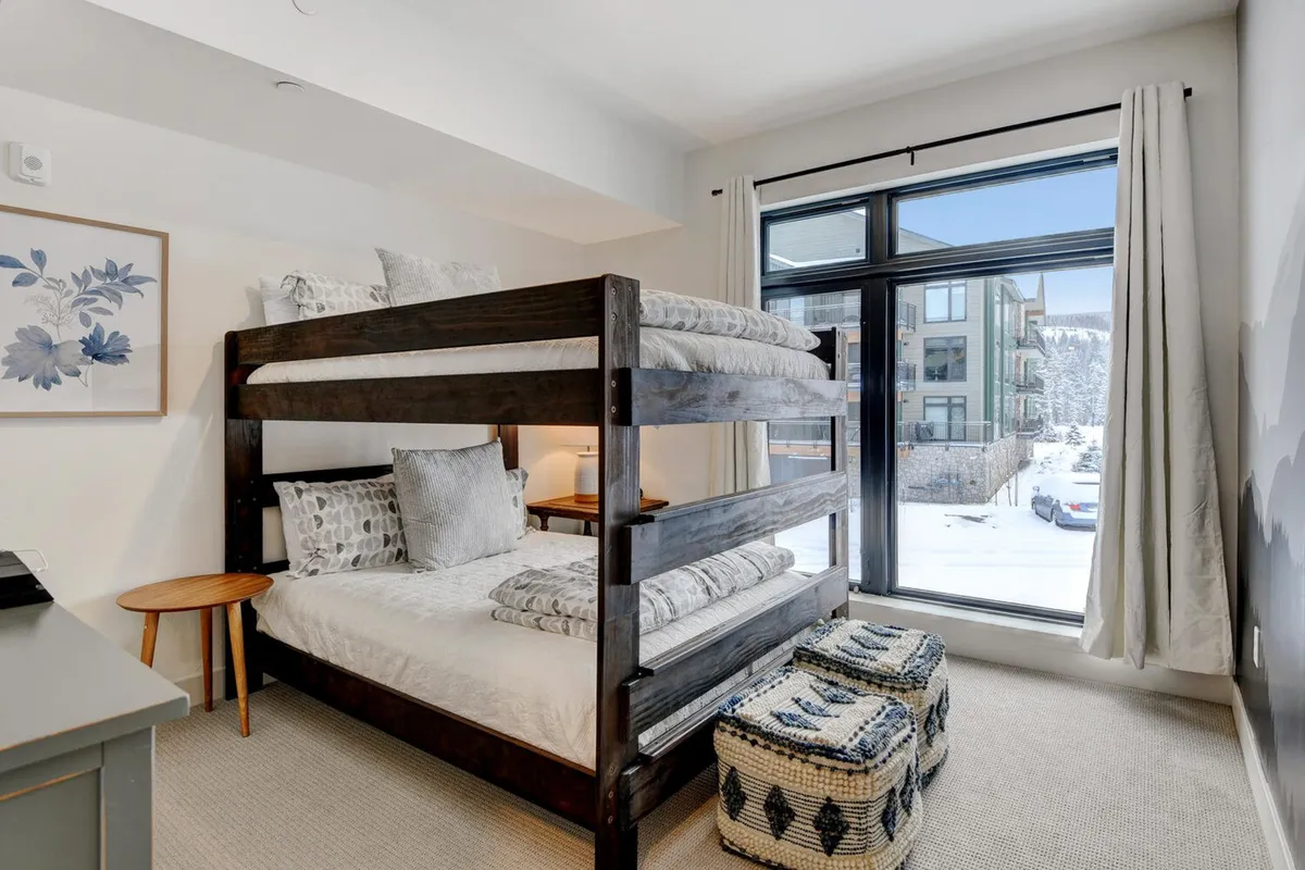 A warm and inviting bunk room with a queen-over-queen bunk bed, large window letting in natural light and snowy views, decorative artwork, and a side table. The space is designed to be comfortable and appealing for both children and adults, perfect for families and groups.