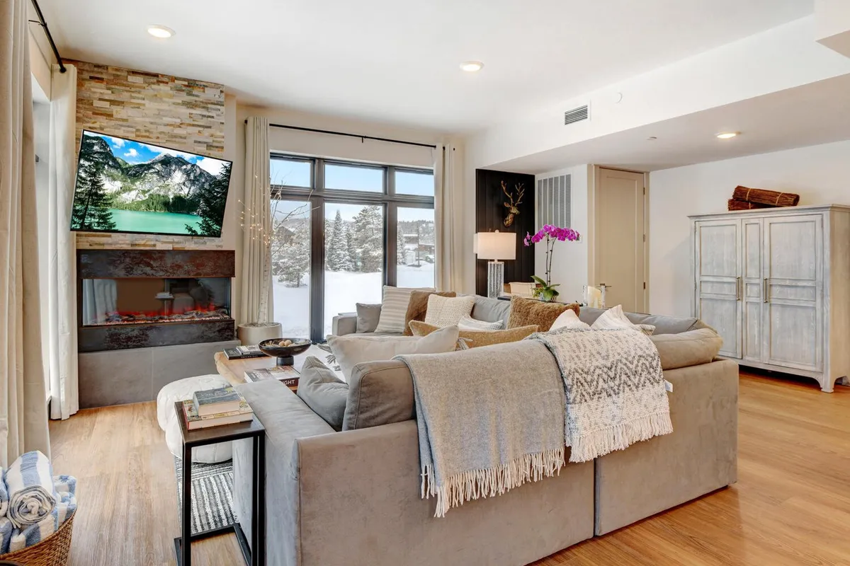 A stylish interior living area with a large sectional sofa, stone accent wall, and a TV positioned to showcase a mountain view. Decorative elements like an orchid and modern lighting add to the upscale, comfortable vibe. The cozy ambiance is enhanced by the suggestion of relaxing by the fireplace, ideal for unwinding after outdoor activities or enjoying time with loved ones.
